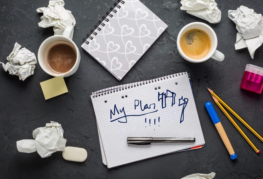 A desk containing scraps of paper and dirty coffee cups, with a pad at the centre with "My Plan" written on it and doodles of axe throwing