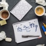 A desk containing scraps of paper and dirty coffee cups, with a pad at the centre with "My Plan" written on it and doodles of axe throwing