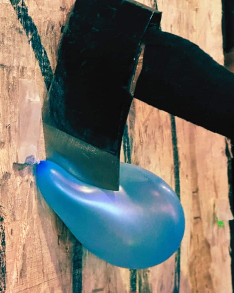 An axe stuck in a wooden target touching but not bursting a balloon on the target